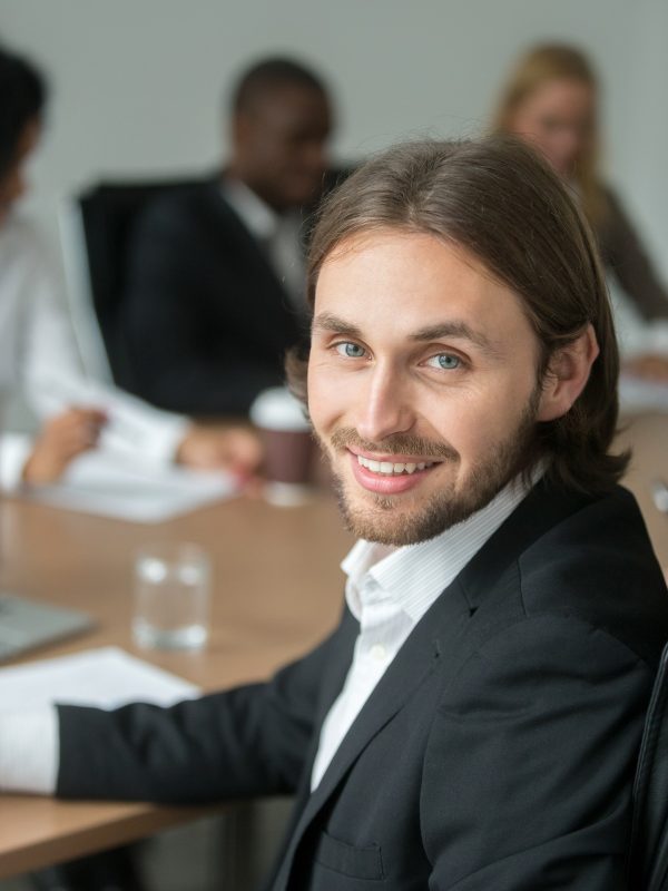 Smiling young businessman in suit looking at camera at meeting, head shot portrait of successful executive team leader, business professional, board member or conference participant, vertical view